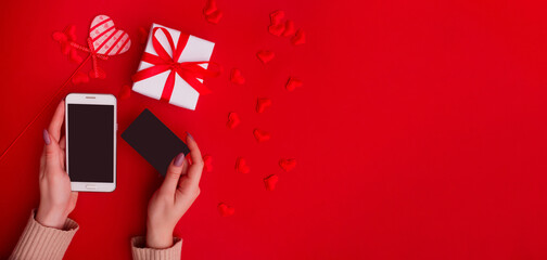 The concept of online shopping, sales on Valentine's Day. Mobile phone and credit card in female hands with gift box and decoration on a red background. Flat lay, top view, copy space, banner.