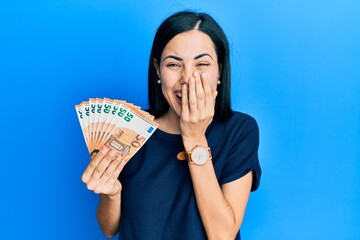 Beautiful young woman holding bunch of 50 euro banknotes laughing and embarrassed giggle covering...