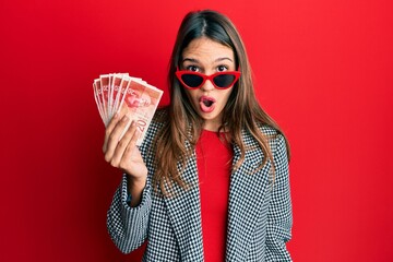 Young brunette woman holding 20 israel shekels banknotes scared and amazed with open mouth for...