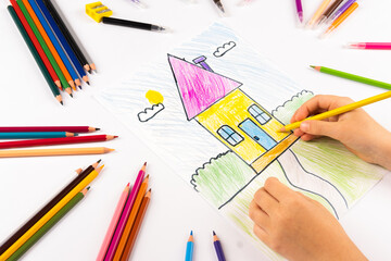 Child girl draws house with multicolored felt-tip pens on a white sheet.