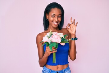 Young african american woman holding flowers doing ok sign with fingers, smiling friendly gesturing excellent symbol