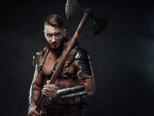 Fototapeta premium Dressed in dark armour with fur dirty and awesome viking fighter with two handed axe on his shoulder poses in dark background.