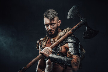 Holding two handed axe on his shoulder scandinavian barbarian in light armour with fur poses in dark background looking at camera. - obrazy, fototapety, plakaty