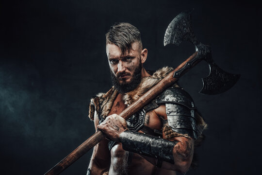 Holding two handed axe on his shoulder scandinavian barbarian in light armour with fur poses in dark background looking at camera.