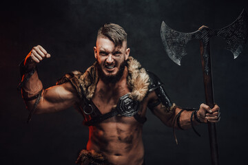 Nordic barbarian with lights armour showing his huge biceps looking at camera and holding his two handed axe in dark background.