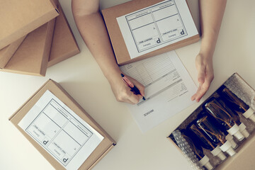 Close-up of woman`s hands on the white desk with carton boxes and signing the parcel with dark glass bottles for delivery. Online working concept.