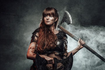 Barbaric female viking in light armour with brown hairs poses in dark smokey background holding two...
