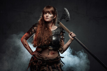 Fototapeta premium Barbaric and brown haired woman viking wielding two handed axe and dressed in dark armour poses in dark smokey background.