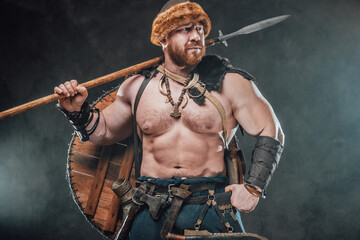 Shirtless and muscular nord barbarian poses in foggy background holding a spear on his shoulder and looking away.