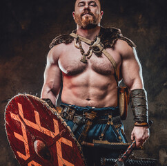Brutal and savage northern barbarian with naked torso and muscular build holding shield and hatchet in dark background.