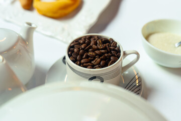Coffee beans in a cup served. White composition with a tea pot, suggar and other elements as a concept of a break. Selective focus.