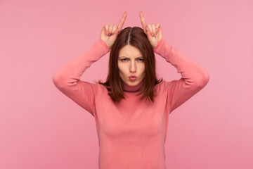 Obraz na płótnie Canvas Arrogant stubborn woman with brown hair in pink sweater showing horns holding fingers above head, demonstrating her aggressive behavior. Indoor studio shot isolated on pink background