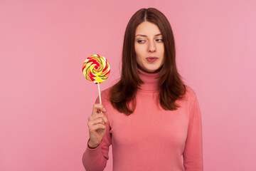 Happy brunette woman in pink sweater looking with desire at striped colorful lollipop on stick in her hand, crazy about sweet confectionery. Indoor studio shot isolated on pink background