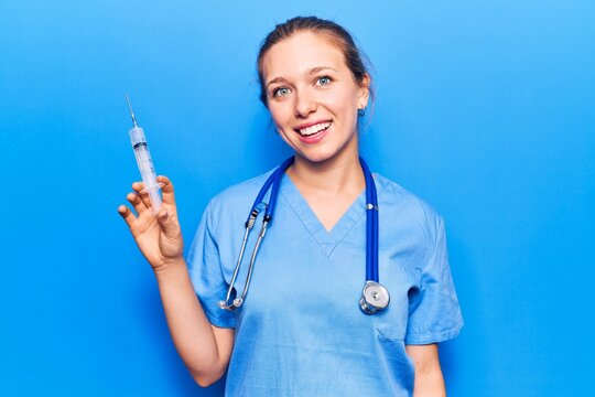 Young blonde woman wearing doctor uniform holding syringe looking positive and happy standing and smiling with a confident smile showing teeth