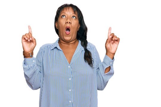 Hispanic middle age woman wearing casual clothes amazed and surprised looking up and pointing with fingers and raised arms.