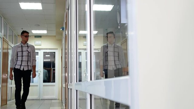 A young man office worker walks along the corridor opens the door and enter it. Reflection in the glass of the guy opening the office door.
