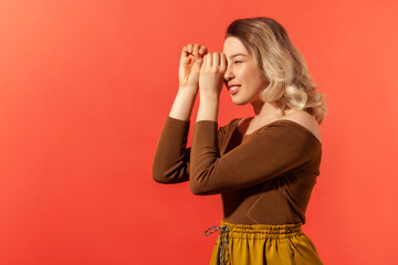 Side view portrait of smiled blonde woman in casual brown blouse making glasses shape, looking through binoculars gesture with surprised expression. Indoor studio shot isolated on red background