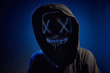 Anonymous man in hood hiding face behind neon glow scary mask on dark background. Horror concept
