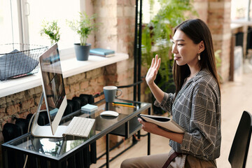 Young female manager in casualwear waving hand to people on computer screen