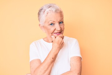 Senior beautiful woman with blue eyes and grey hair wearing classic white tshirt over yellow background looking confident at the camera with smile with crossed arms and hand raised on chin. thinking.