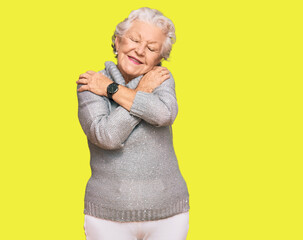Senior grey-haired woman wearing casual winter sweater hugging oneself happy and positive, smiling...