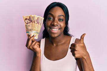 Young african american woman holding hungarian forint banknotes smiling happy and positive, thumb up doing excellent and approval sign