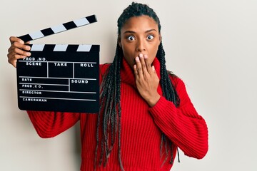 African american woman with braids holding video film clapboard covering mouth with hand, shocked...