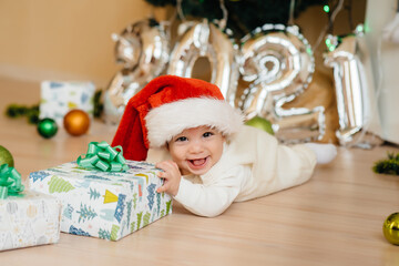 Cute smiling baby is lying under a festive Christmas tree and playing with gifts. Christmas and New Year celebrations