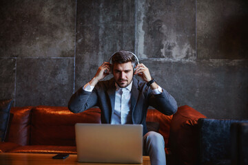 Fototapeta premium Middle-aged man in formal wear sitting in the lobby and putting on earphones. He is looking at the laptop and preparing to have a conference call.