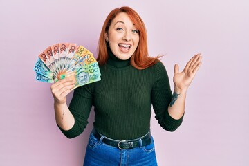 Beautiful redhead woman holding australian dollars celebrating achievement with happy smile and...