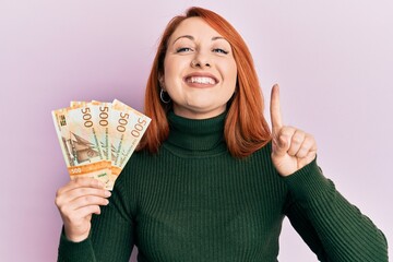 Beautiful redhead woman holding 500 norwegian krone banknotes smiling with an idea or question...
