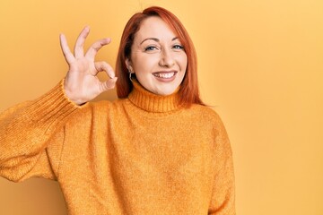 Beautiful redhead woman wearing casual winter sweater over yellow background smiling positive doing...