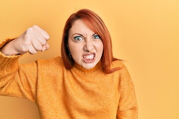 Beautiful redhead woman wearing casual winter sweater over yellow background angry and mad raising...