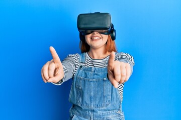 Young redhead woman using 3d virtual glasses standing over isolated blue background.