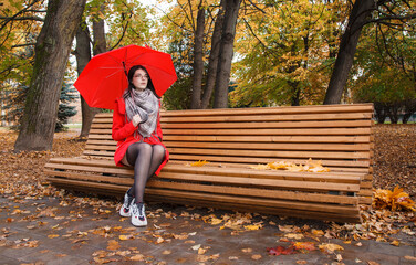 young girl in a red coat with an umbrella sitting on a bench in a city park