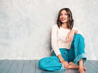 Young beautiful smiling woman looking at camera. Trendy female in casual summer jeans clothes. Positive female with red lips shows facial emotions. Cheerful model sitting near gray wall in studio