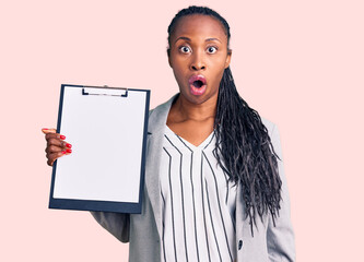Young african american woman wearing business clothes holding clipboard scared and amazed with open...