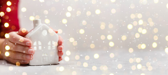 Women hands hold a new small ceramic house with windows with copy space. Golden lights holiday...
