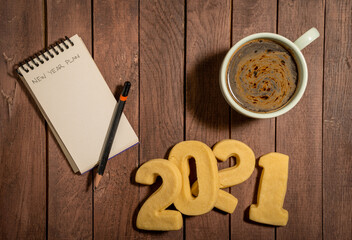 Written New Year Plan text on a notebook with pencil, coffee and cookies on wooden table. Top view, copy space. 2021 new year goals and resolutions concept.