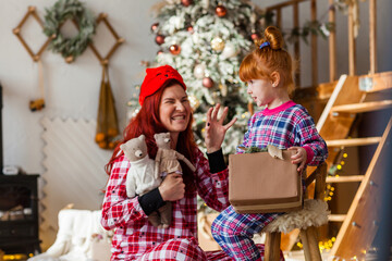 Happy woman and kid wearing checkered pajamas is sitting beside the Christmas tree open presents, winter holidays for mother and daughter