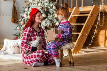 Happy woman and kid wearing checkered pajamas is sitting beside the Christmas tree open presents, winter holidays for mother and daughter