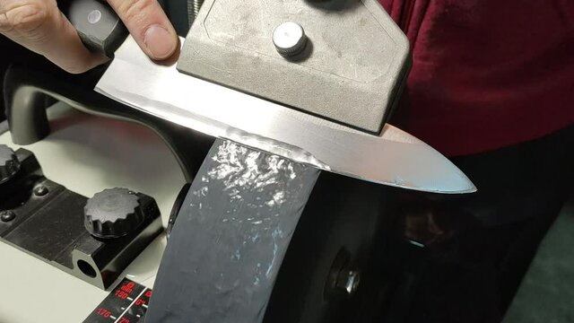 A man sharpens a chef's knife on an electric water grinder on a coarse gray stone.