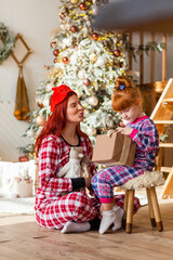 Happy woman and kid wearing checkered pajamas is sitting beside the Christmas tree open presents, winter holidays for mother and daughter