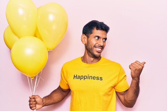 Young latin man wearing t shirt with happiness message holding balloons pointing thumb up to the side smiling happy with open mouth