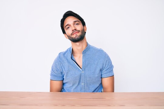 Handsome hispanic man wearing casual clothes sitting on the table relaxed with serious expression on face. simple and natural looking at the camera.