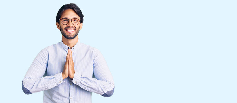 Handsome hispanic man wearing business shirt and glasses praying with hands together asking for forgiveness smiling confident.