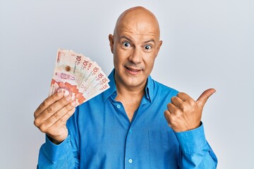 Middle age bald man holding 10 colombian pesos banknotes pointing thumb up to the side smiling happy with open mouth