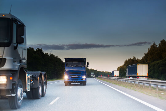 A Group Of Trucks With A Blue Truck In The Center Drive Along A Gray Highway In Summer