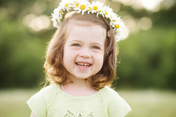 Laughing baby girl 2-3 year old with blonde hair wearing chamomile flower wreath over nature background closeup. Spring season. Childhood. Happiness.