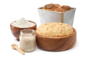 Natural leaven for bread in a glass jar, wooden bowl of dough, bowl of flour and freshly baked loaf in baking tin on white.